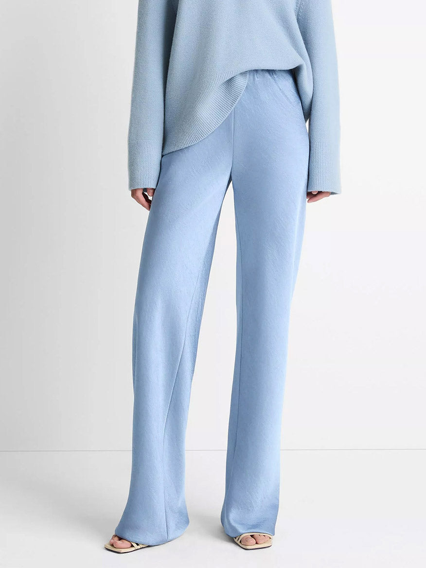 A person stands against a plain background wearing the Vince Fluid Satin Bias Pant in Pacific, styled with an oversized light blue sweater and open-toe shoes.