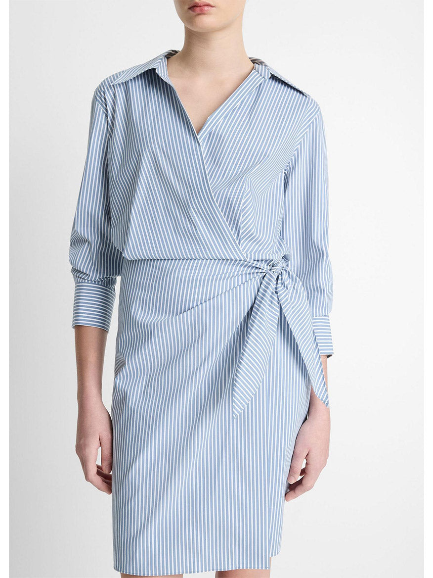 A person stands against a plain white backdrop, showcasing the Vince Furrow Stripe Cotton-Blend Wrap Dress in Coastal Blue/Off-White. The dress features a collar and three-quarter sleeves, crafted from a soft cotton blend for ultimate comfort.