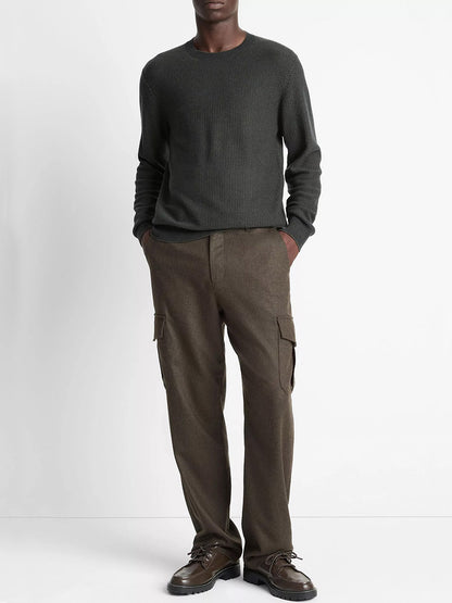A person stands against a plain background wearing the Vince Geo-Jacquard Wool-Blend Crew Neck Sweater in Driver Green/Coastal Blue, brown cargo pants, and dark shoes, with hands in their pockets.