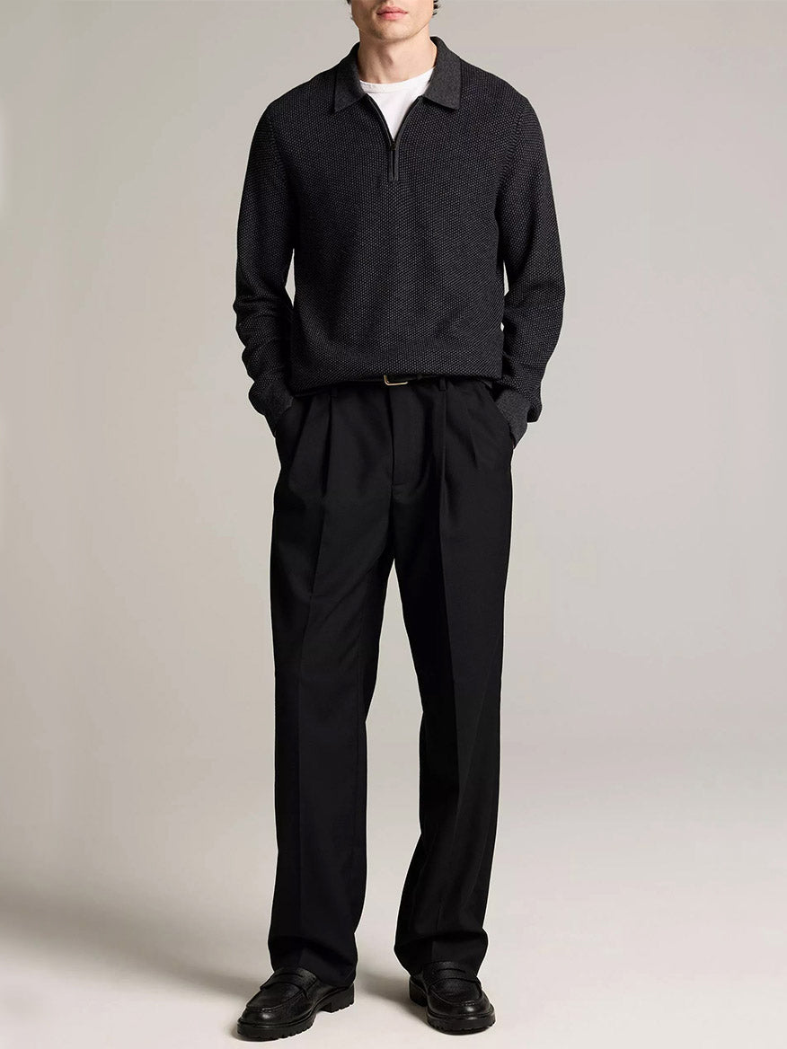 A person stands with hands in pockets, wearing the Vince Geo-Jacquard Wool-Blend Zip Polo Sweater in Coastal Blue/Medium Heather Grey over a white shirt, paired with black pleated trousers and black shoes, against a plain light background.