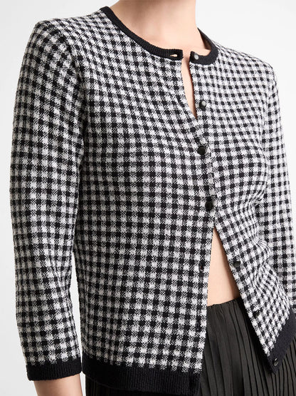 A person wears the Vince Gingham Italian Wool Blend Cardigan in Black/Off-White, featuring black buttons and three-quarter sleeves, paired with a pleated black skirt.