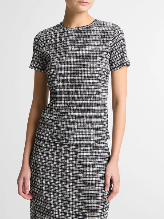 A woman wears the Vince Plaid Grid-Knit Short-Sleeve Crew Neck Top in Eclipse Combo, featuring a black-and-white plaid pattern with stretchy smocking, paired with a matching skirt against a light background.