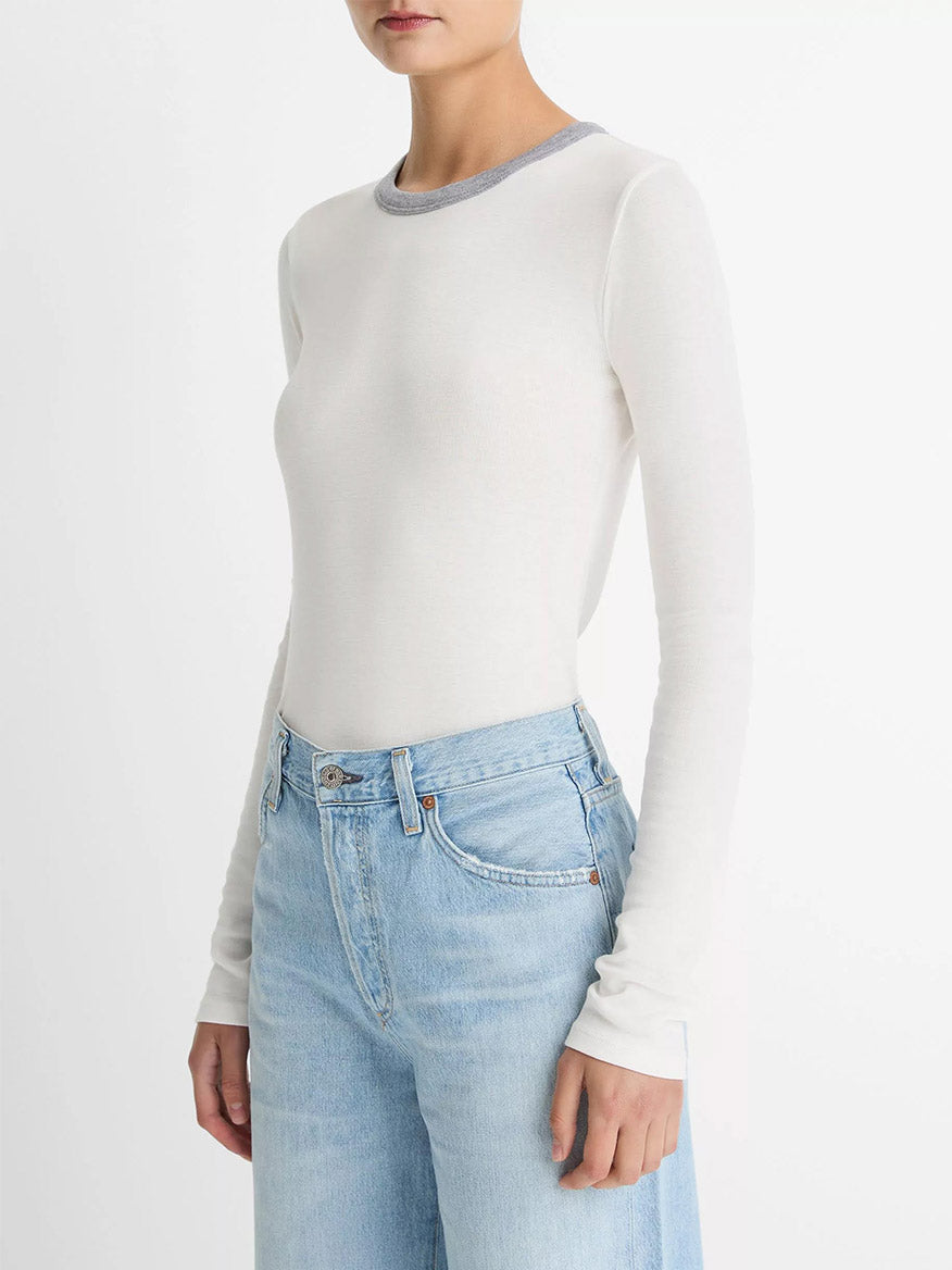 Wearing the Vince Tipped Heathered Pima Cotton-Blend Long-Sleeve T-Shirt in Off White Combo and high-waisted light blue jeans, a person stands against a plain white background.