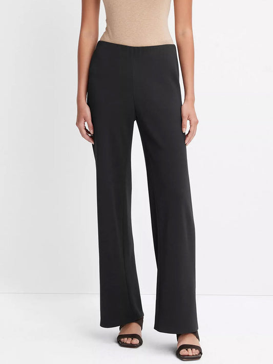 A person stands against a plain white background, wearing a beige sleeveless top with Vince High-Waist Crepe Bias Pant in Black and black sandals.