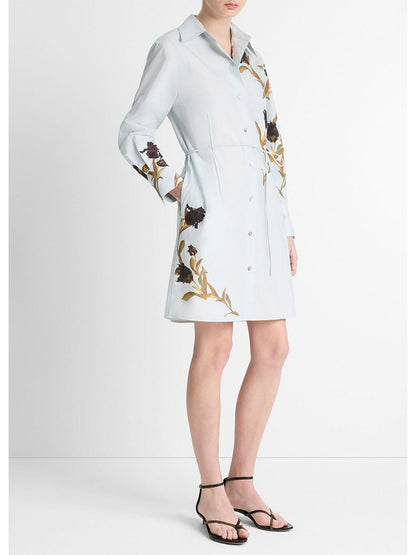 A person in a Vince Placed Iris Cotton Shirt Dress in Salt Glass/Black Iris, paired with black sandals and featuring a chic self-tie belt, stands against a plain background.