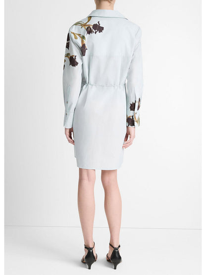 The Vince Placed Iris Cotton Shirt Dress in Salt Glass/Black Iris showcases its elegant design, featuring dark irises on light blue cotton poplin. This knee-length shirt dress includes long sleeves and a self-tie belt, viewed from the back with black heels to enhance the stylish look.