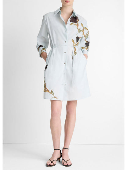 Wearing the Vince Placed Iris Cotton Shirt Dress in Salt Glass/Black Iris, a person is adorned with delicate iris patterns. The self-tie belt cinches the waist elegantly, and hands rest in pockets complementing the look with black sandals.