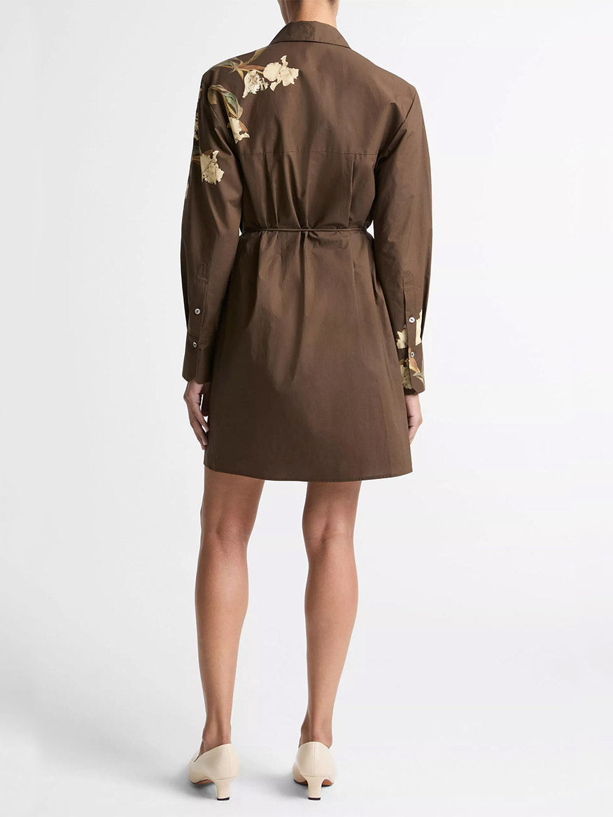 A person wearing the Vince Placed Iris Cotton Shirt Dress in Dark Bark, featuring floral embroidery and a self-tie belt, seen from the back. They are also wearing light-colored shoes.