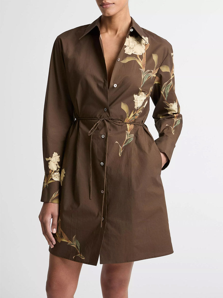 A person wears the Vince Placed Iris Cotton Shirt Dress in Dark Bark, featuring a self-tie belt and yellow iris floral print on the front and sleeves.