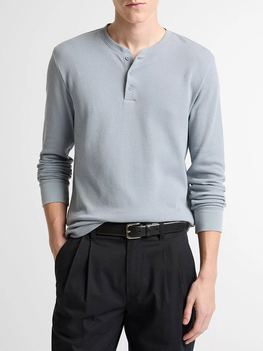 A person wearing a Vince Jacquard-Rib Cotton Henley in Steel Blue and black pleated pants with a black belt, standing with one hand in their pocket.