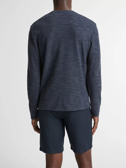A person stands facing away, wearing the Vince Double-Knit Cotton-Blend Long-Sleeve Shirt in Heather Coastal Blue and dark shorts against a plain light background. The shirt offers sweater-like softness for ultimate comfort.