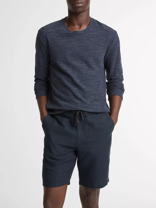 A person wears the Vince Double-Knit Cotton-Blend Long-Sleeve Shirt in Heather Coastal Blue with dark drawstring shorts, standing with hands in pockets against a plain background.