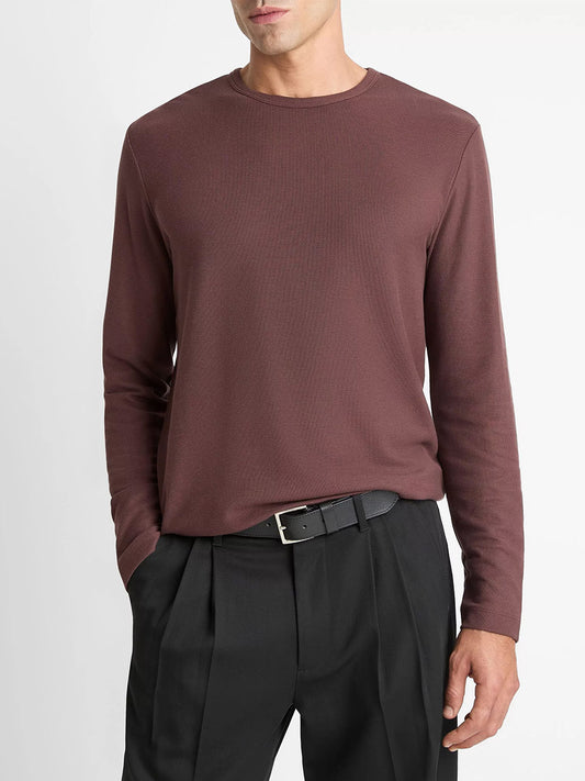 A man wears a Vince Pima Cotton Piqué Long-Sleeve T-Shirt in Washed Port, tucked into pleated black trousers with a black belt, standing against a plain white background.