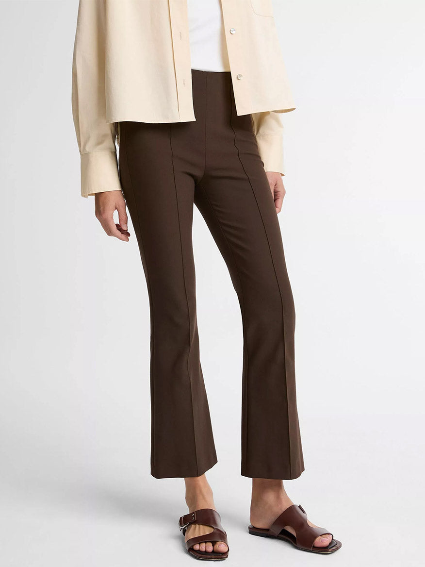 A woman wears the Vince Niki Mid-Rise Pintuck Crop Flare Pant in Willow, paired with a cream button-up shirt, white top, and brown slide sandals, standing against a plain white background.
