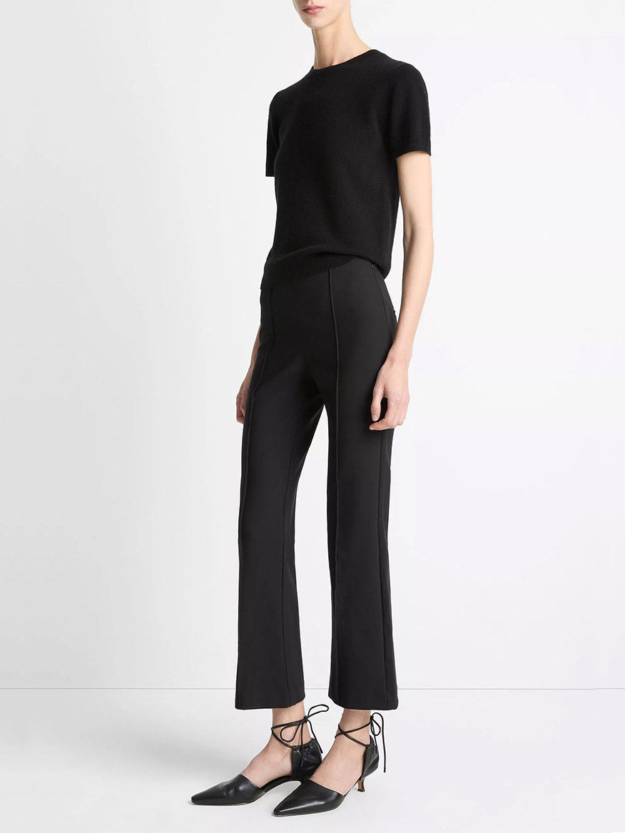 A person wearing a short-sleeve black top, Vince Mid-Rise Pintuck Crop Flare Pant in Black, and black pointed-toe lace-up heels stands against a plain white background.