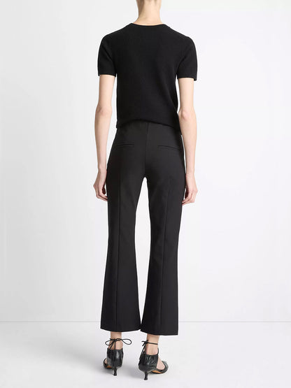 A person stands facing away against a white background, wearing a short-sleeve black top, Vince Mid-Rise Pintuck Crop Flare Pant in Black, and black heeled sandals.