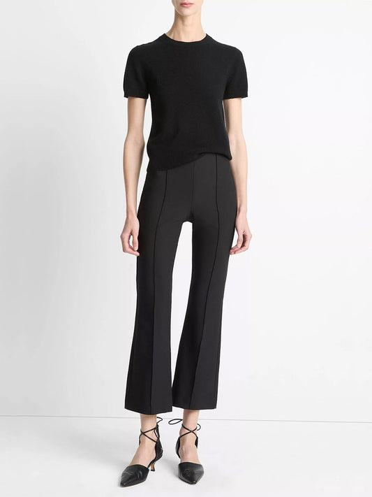 Person standing against a white background, wearing a Vince short-sleeve black top, Vince Mid-Rise Pintuck Crop Flare Pant in Black, and black pointed-toe shoes with ankle ties.