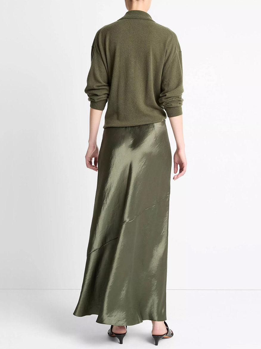 A person stands facing away, wearing an olive green long-sleeve sweater and the Vince Paneled Satin Bias Skirt in Herba with sandals, set against a plain white background.