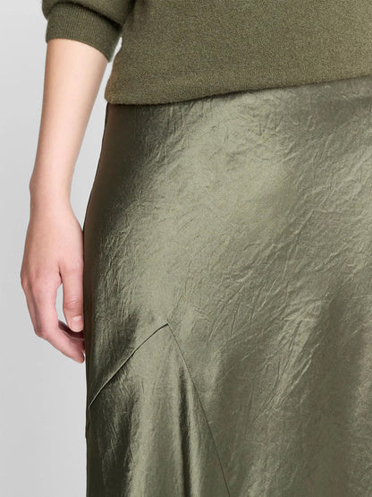 A close-up of a person wearing an olive green sweater and the Vince Paneled Satin Bias Skirt in Herba, featuring shiny, slightly wrinkled Naia acetate satin with an asymmetric design.