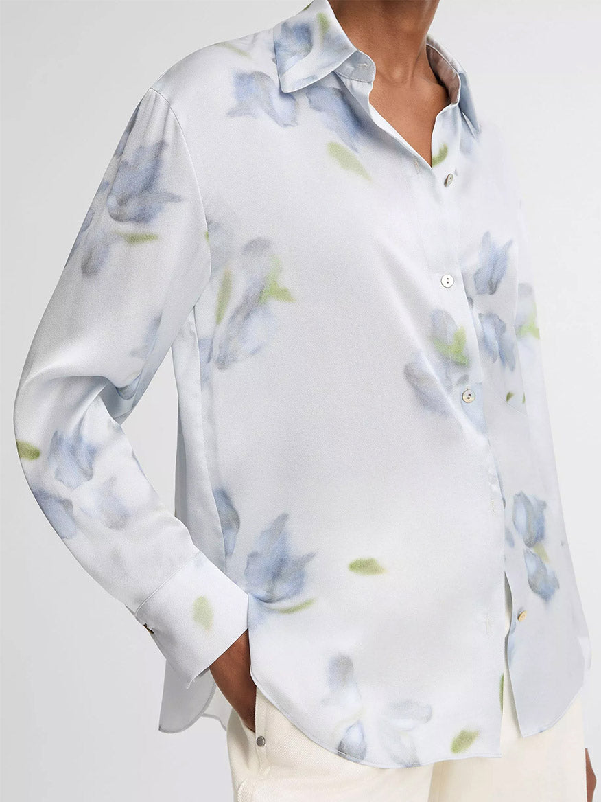 Person wearing the Vince Petal Blur Silk Relaxed Button-Front Shirt in Soft Grey, a long-sleeved white silk shirt with a light blue and green floral pattern and Agoya shell buttons, paired with off-white pants.