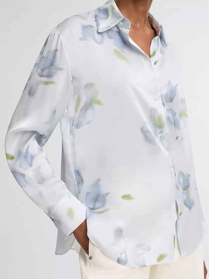 Person wearing the Vince Petal Blur Silk Relaxed Button-Front Shirt in Soft Grey, a long-sleeved white silk shirt with a light blue and green floral pattern and Agoya shell buttons, paired with off-white pants.