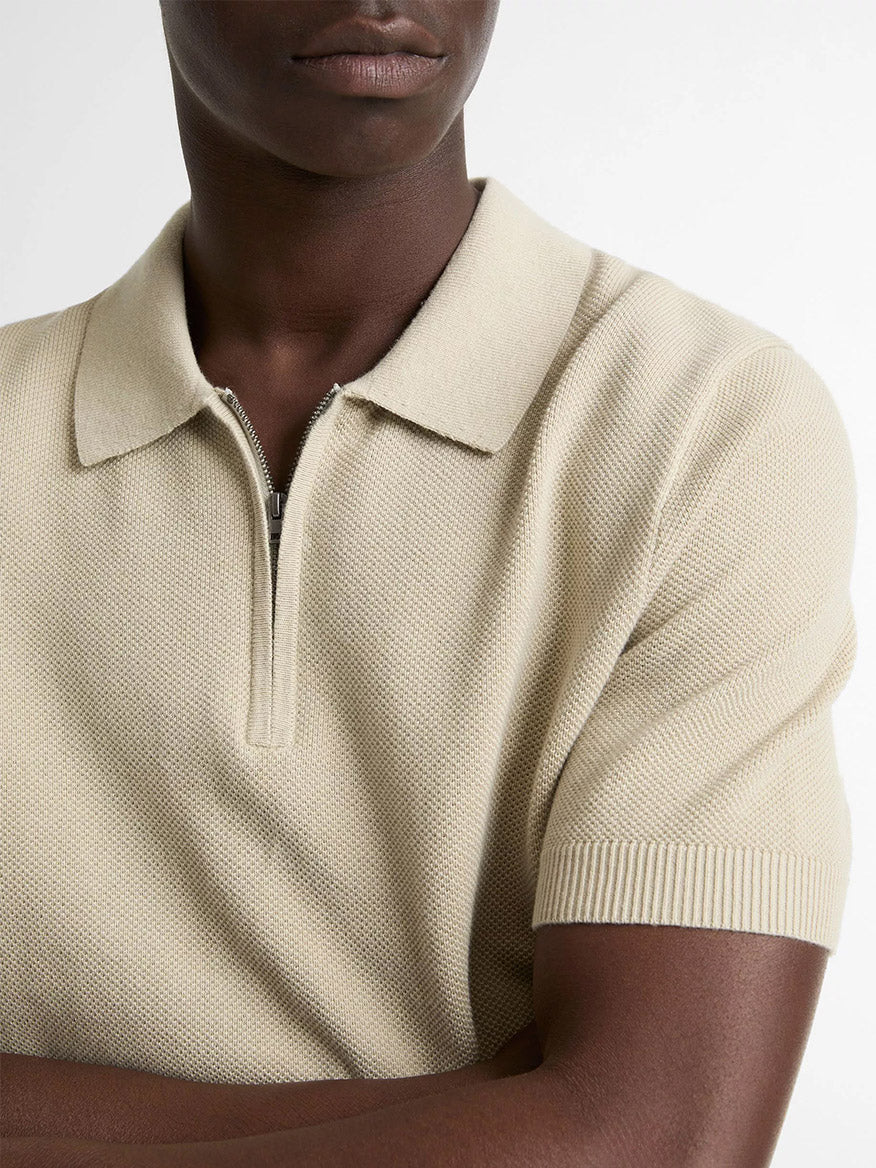 A person wears the Vince Cotton-Cashmere Piqué Zip Polo Sweater in Parchment, featuring short sleeves and a zippered collar, standing with arms crossed.