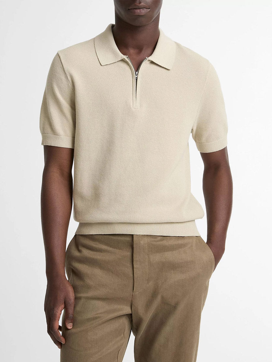 A person wears the Vince Cotton-Cashmere Piqué Zip Polo Sweater in Parchment with brown trousers, standing with one hand in their pocket against a plain backdrop.