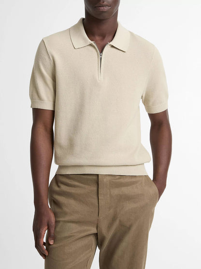 A person wears the Vince Cotton-Cashmere Piqué Zip Polo Sweater in Parchment with brown trousers, standing with one hand in their pocket against a plain backdrop.