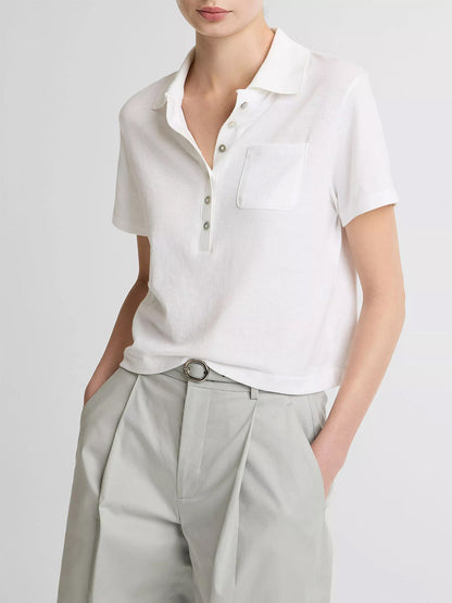 Wearing the Vince Cotton Cropped Pocket Polo in Optic White with agoya shell buttons and a chest pocket, paired with light gray pleated trousers, standing hands in pockets.