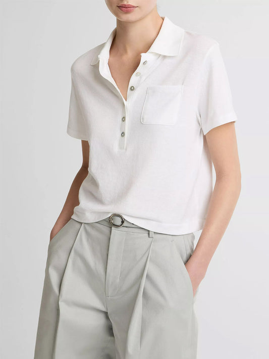 Wearing the Vince Cotton Cropped Pocket Polo in Optic White with agoya shell buttons and a chest pocket, paired with light gray pleated trousers, standing hands in pockets.