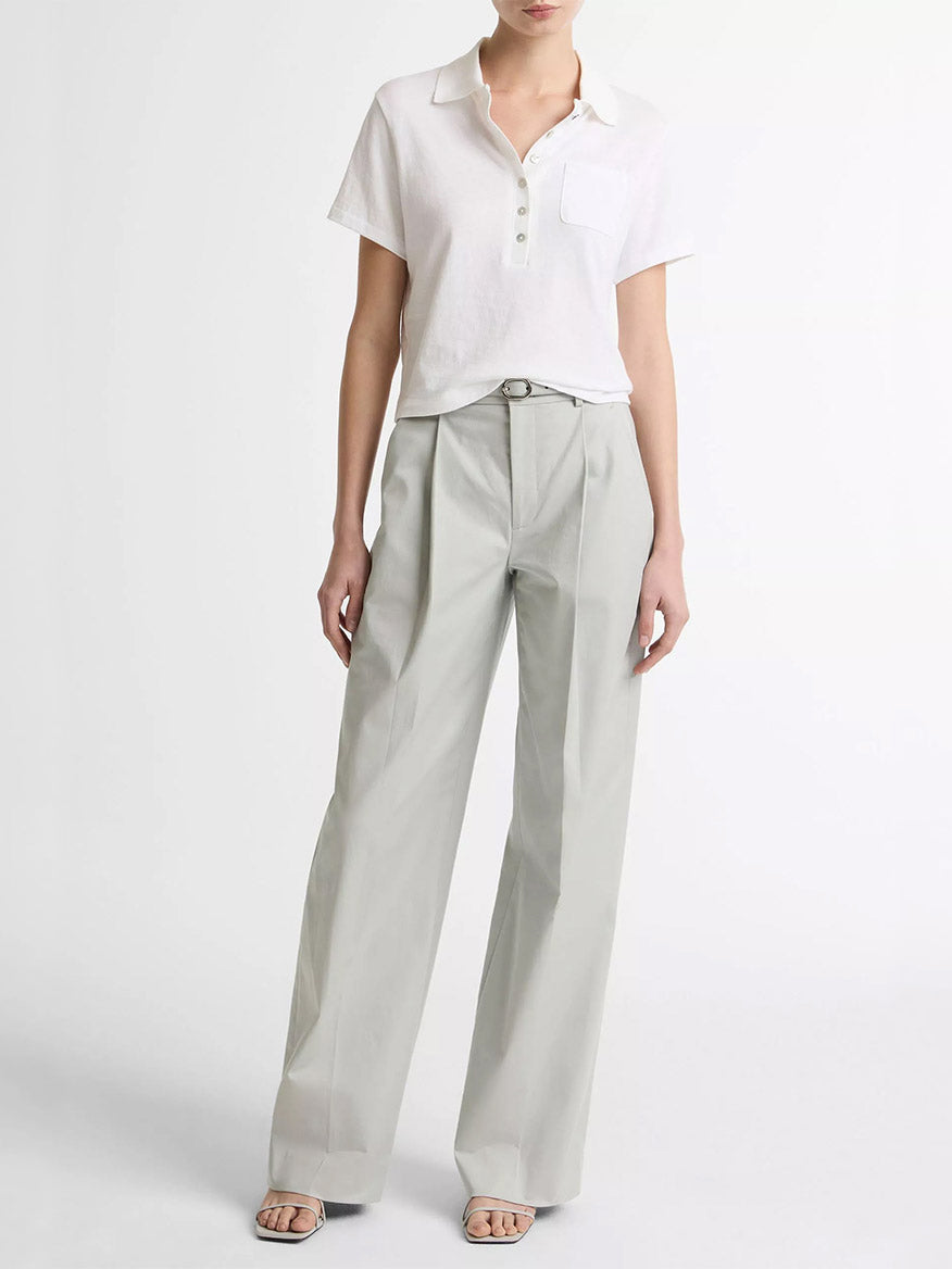 A person wears the Vince Cotton Cropped Pocket Polo in Optic White with light gray high-waisted pleated trousers and open-toe sandals, standing against a plain white background.