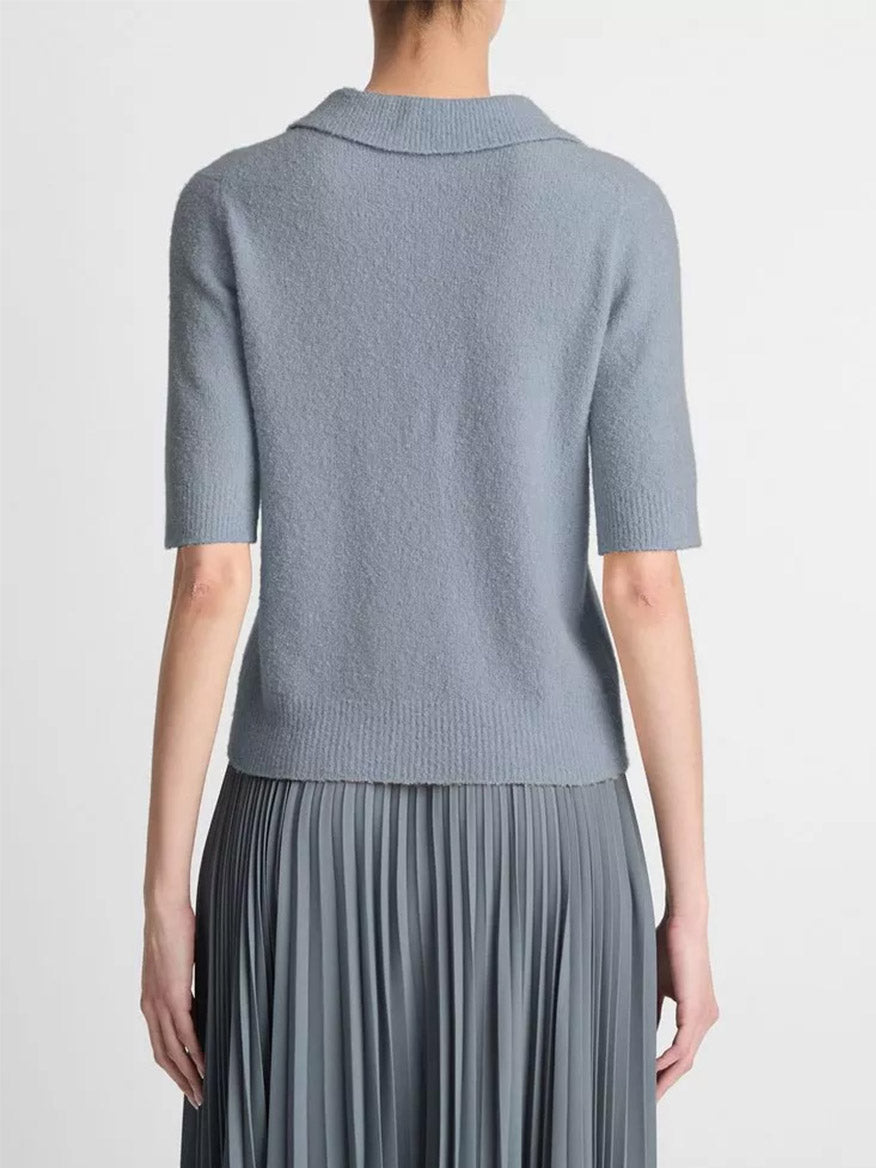 A person wearing a Vince Soft Bouclé Silk-Blend Polo Sweater in Lake Stone and a pleated gray skirt, photographed from the back against a neutral background.