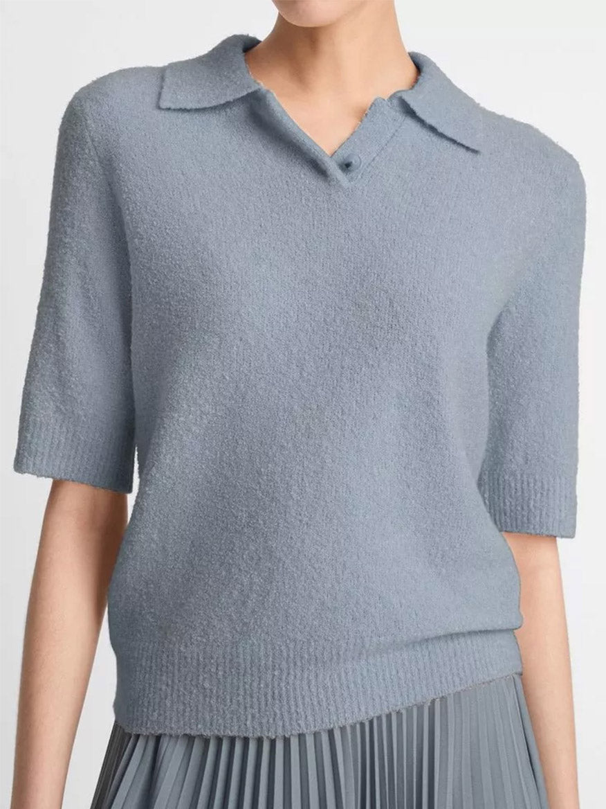 A person wearing the Vince Soft Bouclé Silk-Blend Polo Sweater in Lake Stone, a light blue short-sleeved polo with a collared neckline, paired with a pleated skirt.