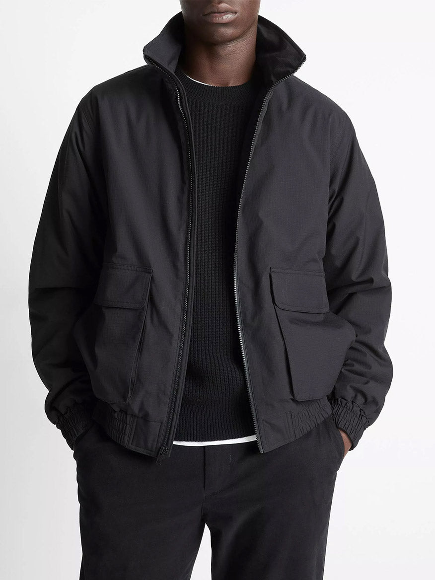 A person wears the Vince Italian Cotton Ripstop Bomber Jacket in Black, paired with a black sweater and black pants, standing hands-in-pockets against a plain background.