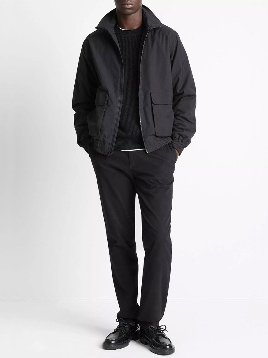 A person stands against a white background wearing the Vince Italian Cotton Ripstop Bomber Jacket in Black, paired with a black sweater, black pants, and black shoes, with hands in the jacket pockets.