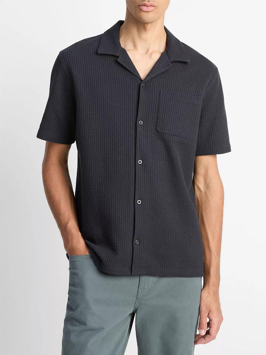 A man wears the Vince Seersucker-Knit Cotton Cabana Shirt in Coastal Blue with a button-front closure and chest pocket, paired with light gray pants, standing against a plain light background.