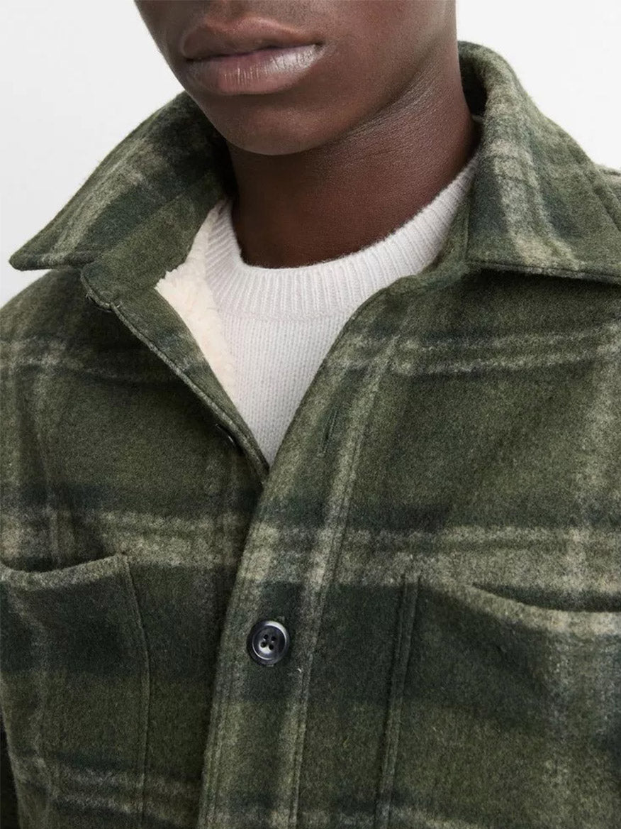 Close-up of a person wearing the Vince Sherpa-Lined Plaid Shirt Jacket in Night Moss Combo with button-front closure over a white sweater. Only the lower half of their face and chest are visible.