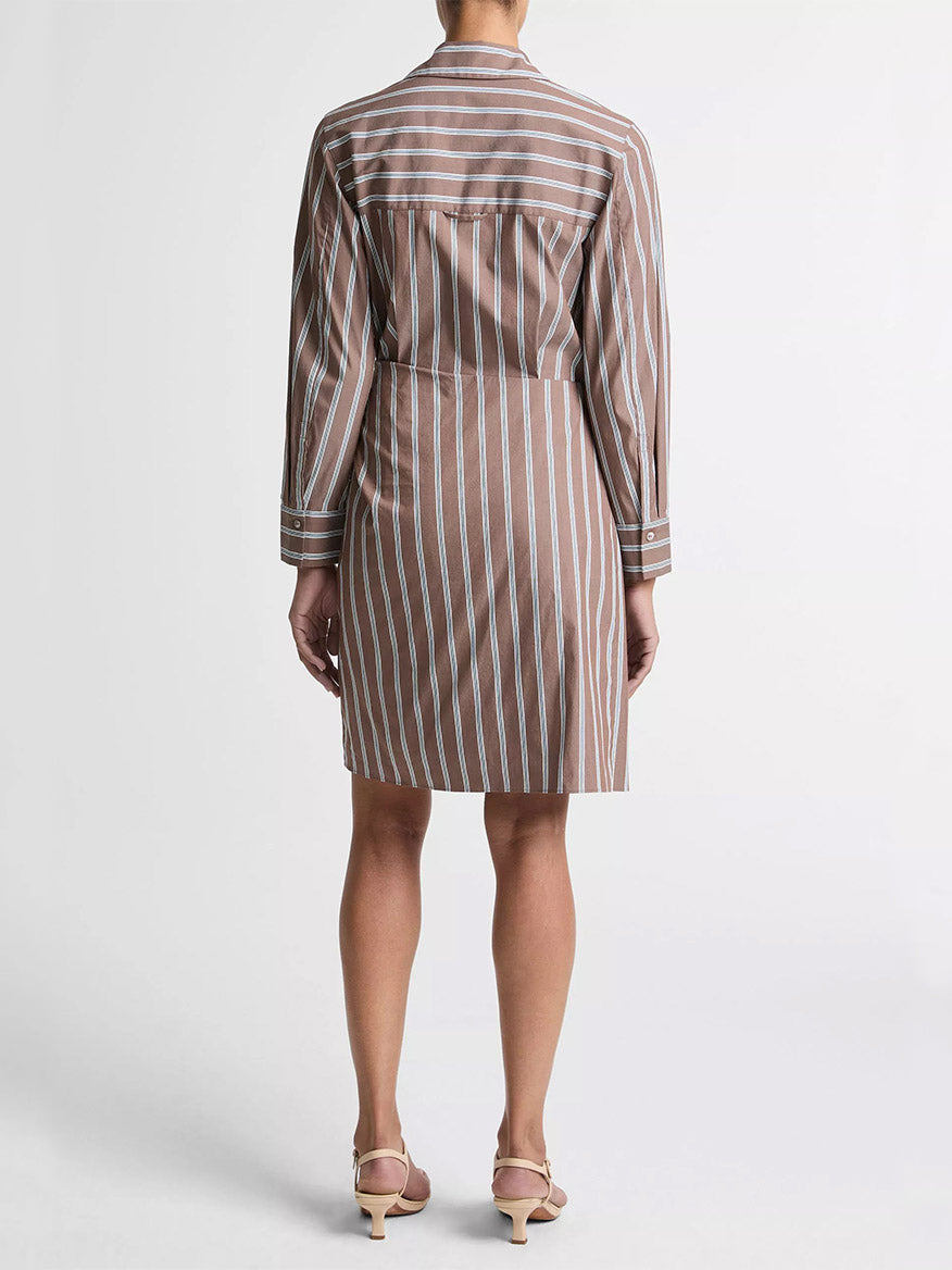 A person stands facing away, wearing the Vince Striped Cotton-Blend Wrap Dress in Brown Multi with long sleeves and beige heeled sandals against a plain white background.