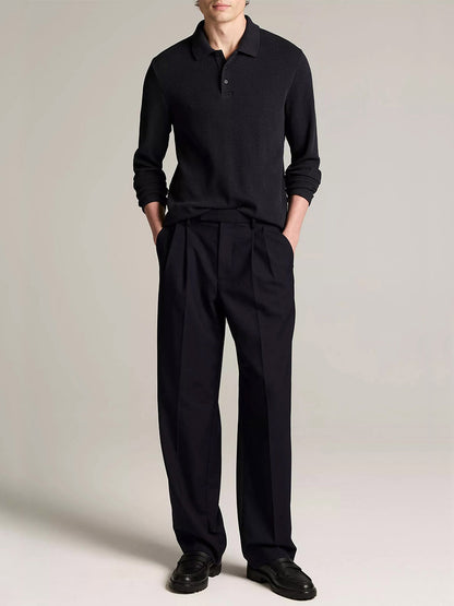 A man stands against a plain background in the Vince Variegated Cotton Polo Shirt in Coastal Blue, dark pleated trousers, and black shoes, hands in pockets, face partially out of frame.
