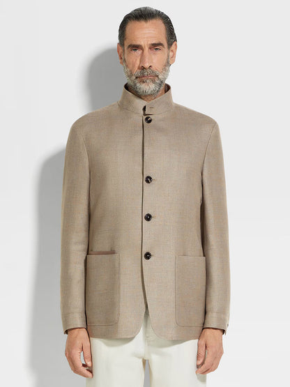 A man with gray hair and beard wears a Zegna Il Conte Chore Jacket in Light Taupe, featuring dark buttons and large front pockets, paired with white trousers as he stands against a plain background.