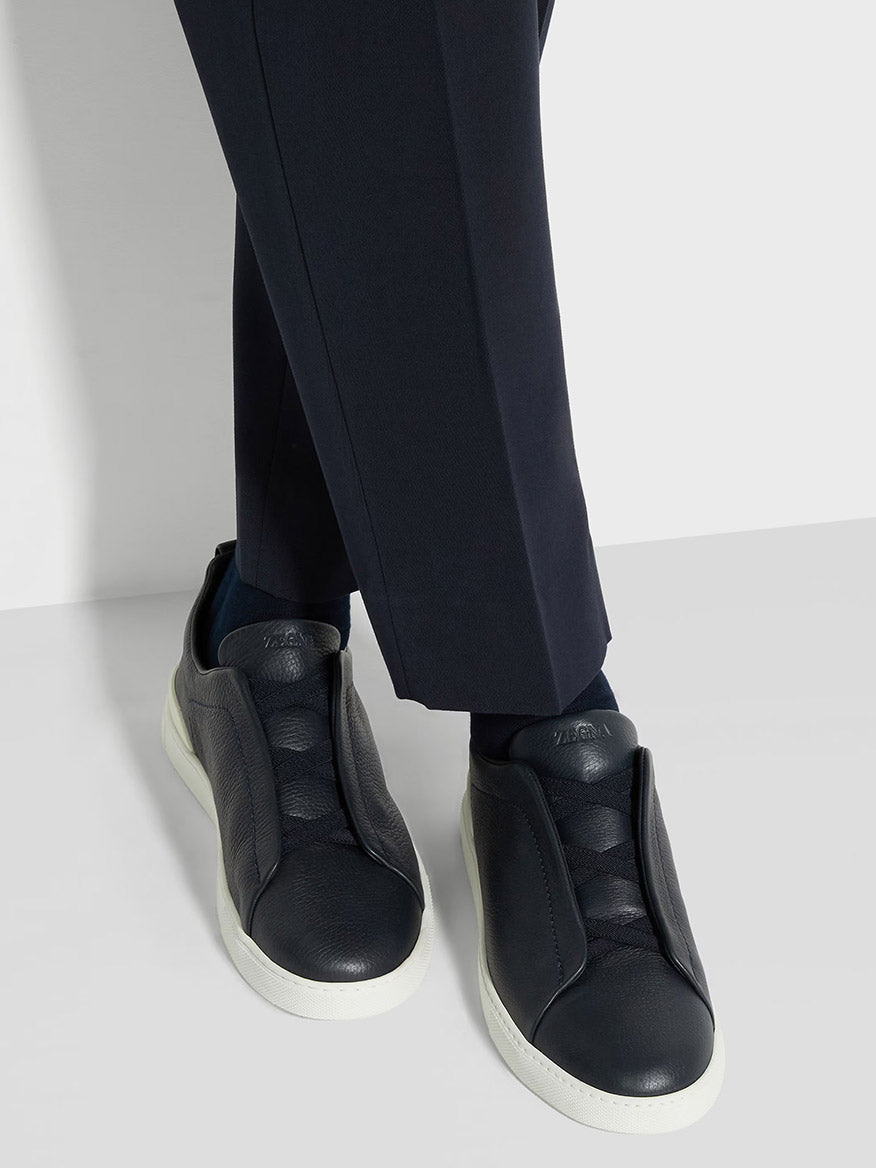 A person wearing sleek Zegna Navy Deerskin Triple Stitch™ Sneakers with white soles and black pants stands against a minimal white background.