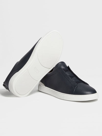 A pair of navy blue Zegna Triple Stitch™ sneakers in premium deerskin, featuring a slip-on low-top design with white rubber soles—one upright, the other showing the sole.