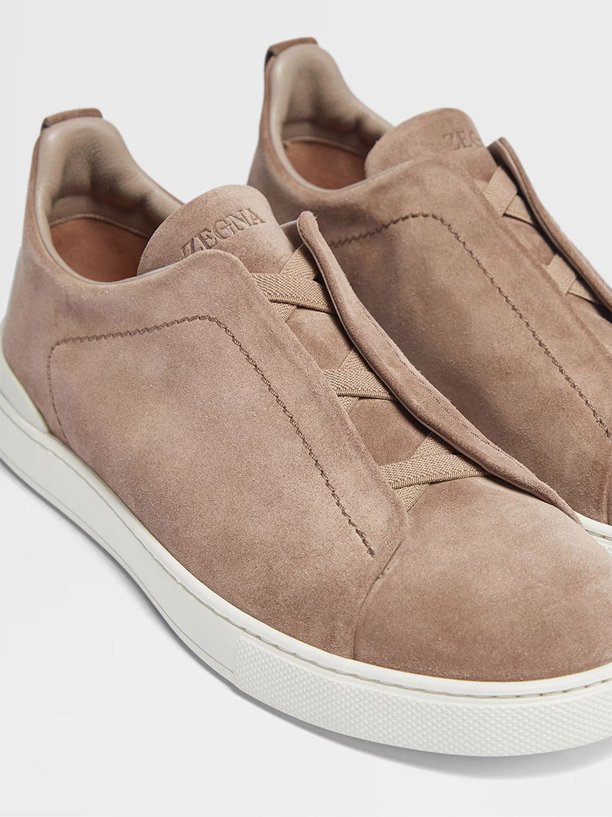 The Zegna Beige Suede Triple Stitch Low Top Sneaker boasts a sleek design with beige suede material and white rubber soles. It features concealed laces and "Zegna" branding on the tongue and back, complemented by their signature Triple Stitch detailing.