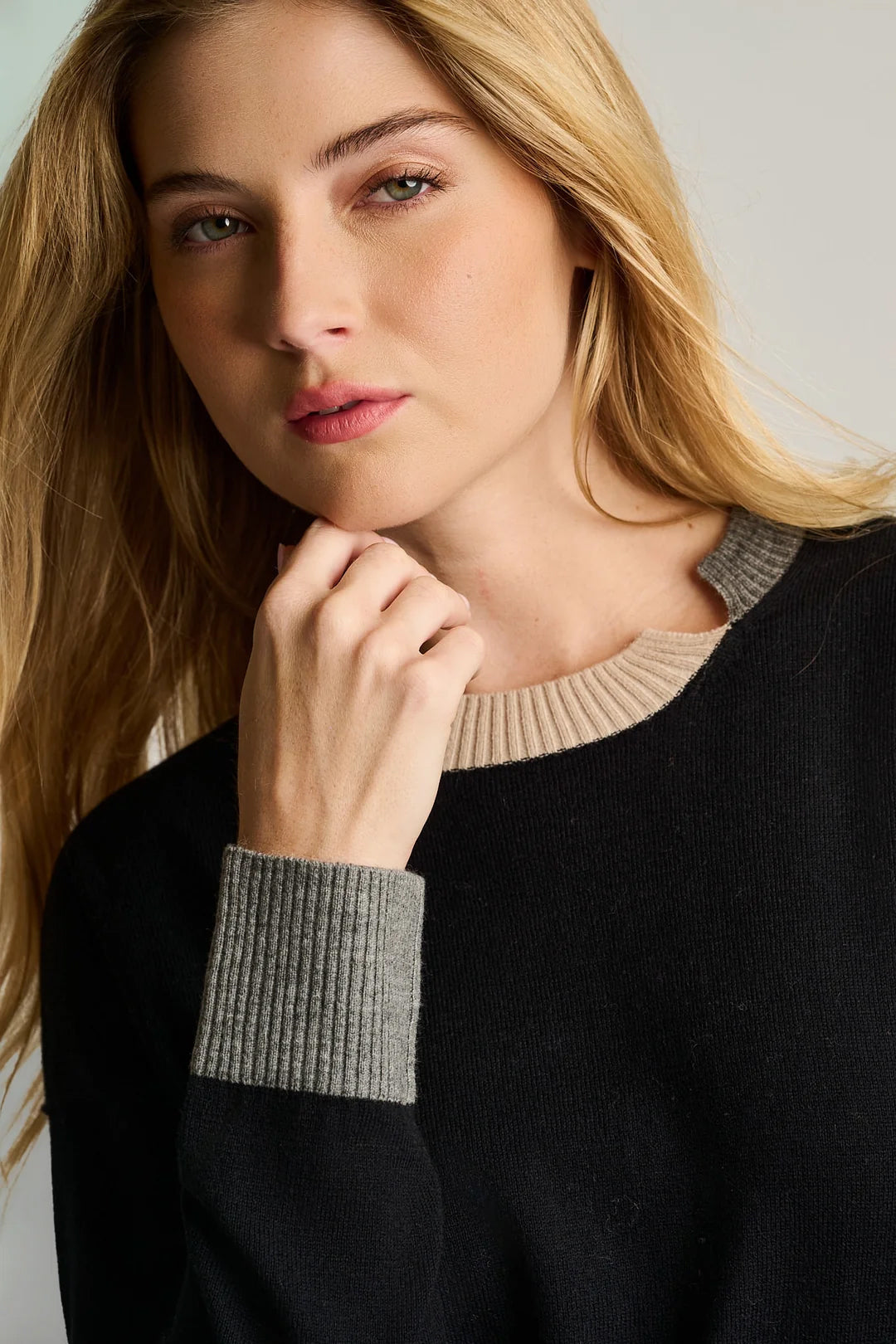 A woman with long blonde hair wears the Lisa Todd Bold Block Cotton Cashmere Sweater in black, featuring a beige collar and gray cuffs, as she looks at the camera with her hand near her chin.