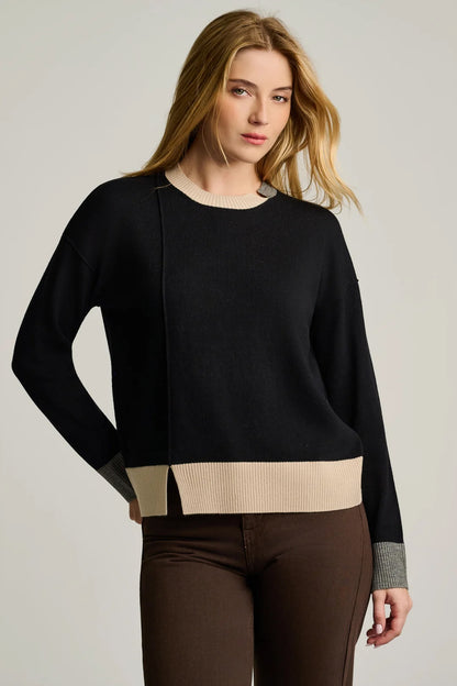 A woman stands against a plain background wearing the Lisa Todd Bold Block Cotton Cashmere Sweater in Black—a lightweight knit sweater with a black body, beige collar and hem, gray cuffs—paired with brown pants.