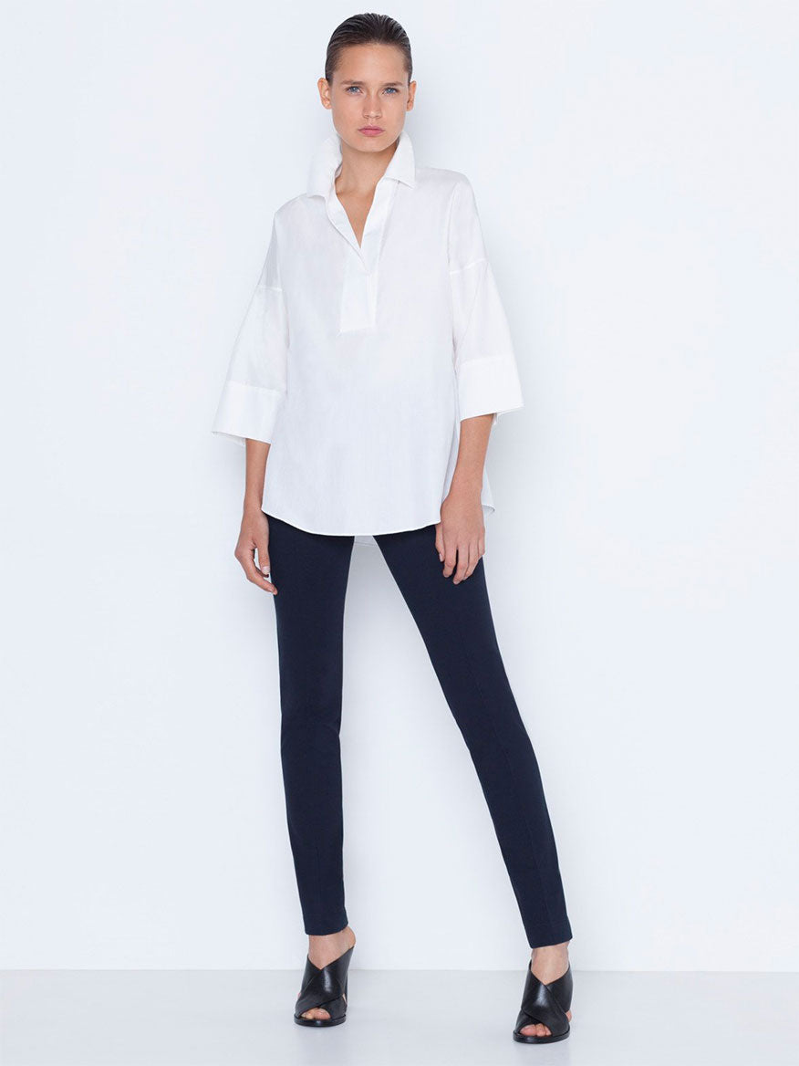 A woman stands against a white background wearing a white oversized shirt paired with Akris Punto Mara Stretch Jersey Pants in Navy and black open-toe heels.