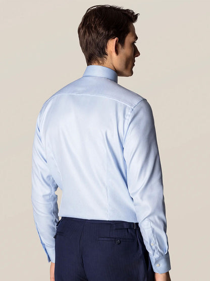 A man showcases refined business attire, standing with his back to the camera in an Eton Slim Fit Light Blue Textured Twill Dress Shirt and dark trousers against a plain background.