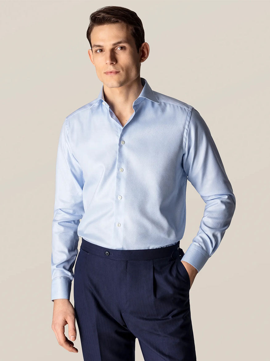 A man in an Eton Slim Fit Light Blue Textured Twill Dress Shirt and dark dress pants stands against a plain, light background with one hand in his pocket, exemplifying polished business attire.
