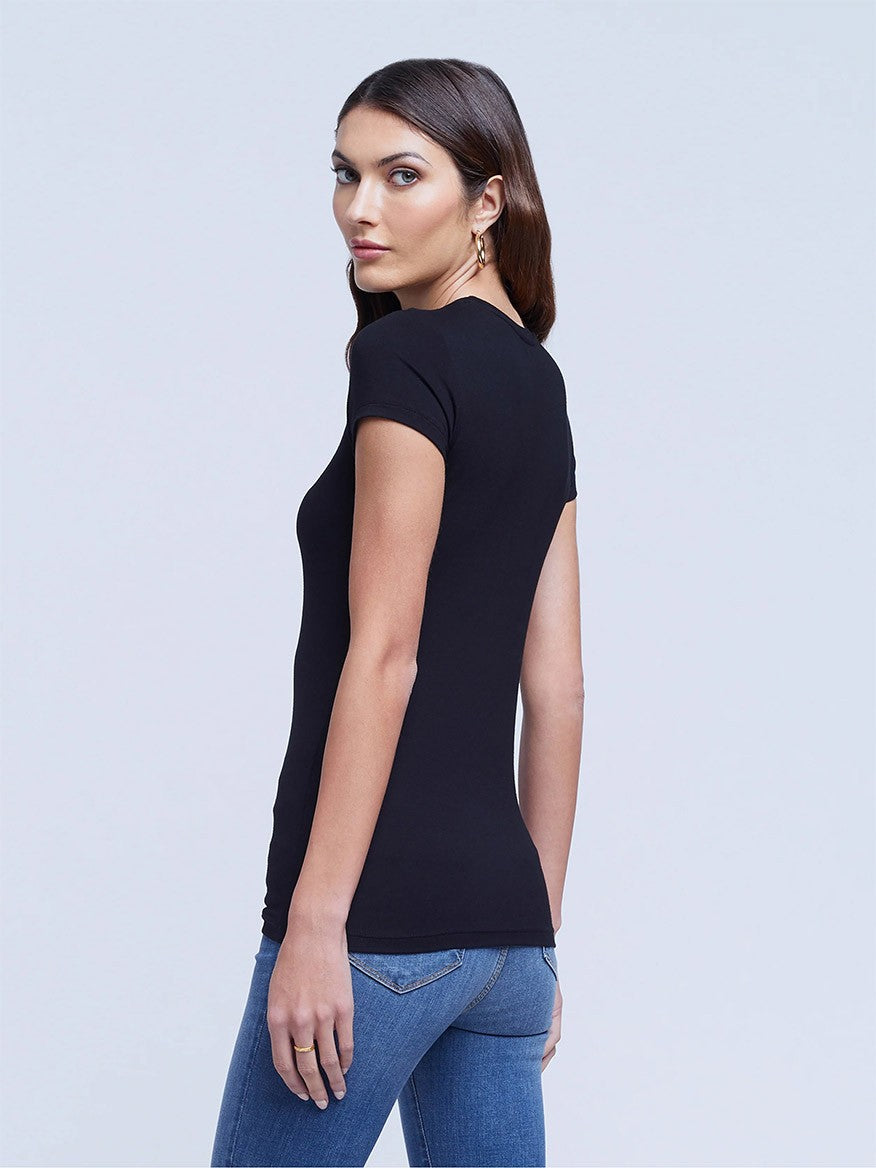 A woman in the L'Agence Ressi Short Sleeve Crew in Black and blue jeans stands turned slightly, looking over her shoulder against a light gray background.