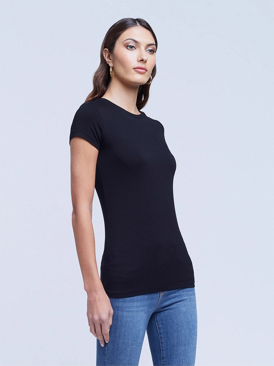 Woman in a L'Agence Ressi Short Sleeve Crew in Black and blue jeans standing with a slight turn to her side against a gray background.